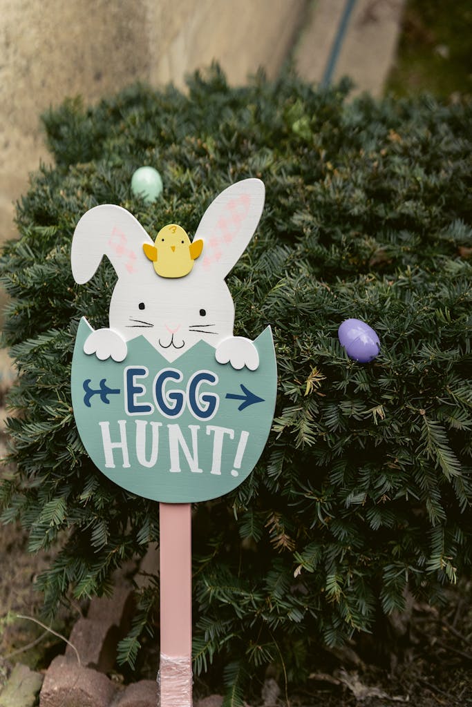 Decorative sign with inscription and bunny placed near green branches of tree with colorful eggs during Easter celebration on street